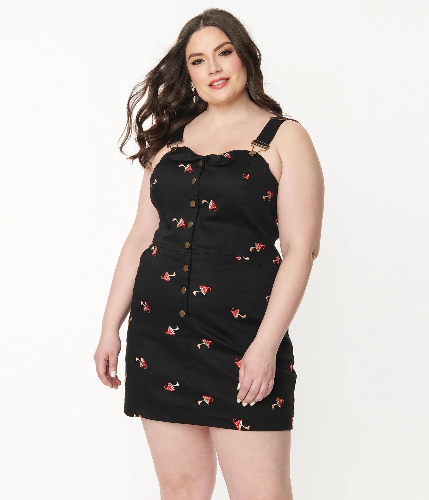 Fair skinned pretty woman standing with long beach wave hair wearing black denim pinafore dress with mushrooms embroidered all over.