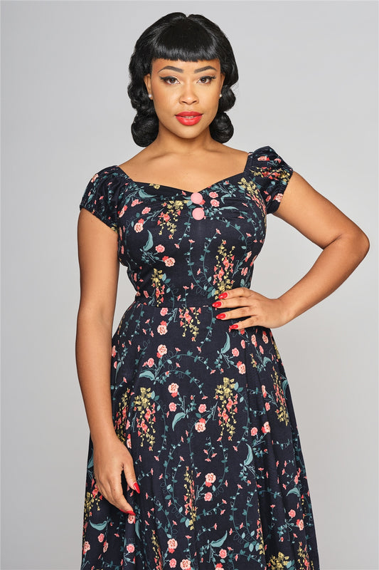 Glamorous woman with short, curled blackhair and red lipstick standing with one hand on her hip in a floral print navy blue 50s style dress with short sleeves.