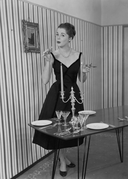 A black and white photo of a 50s housewife wearing a black evening dress preparing the dinner table