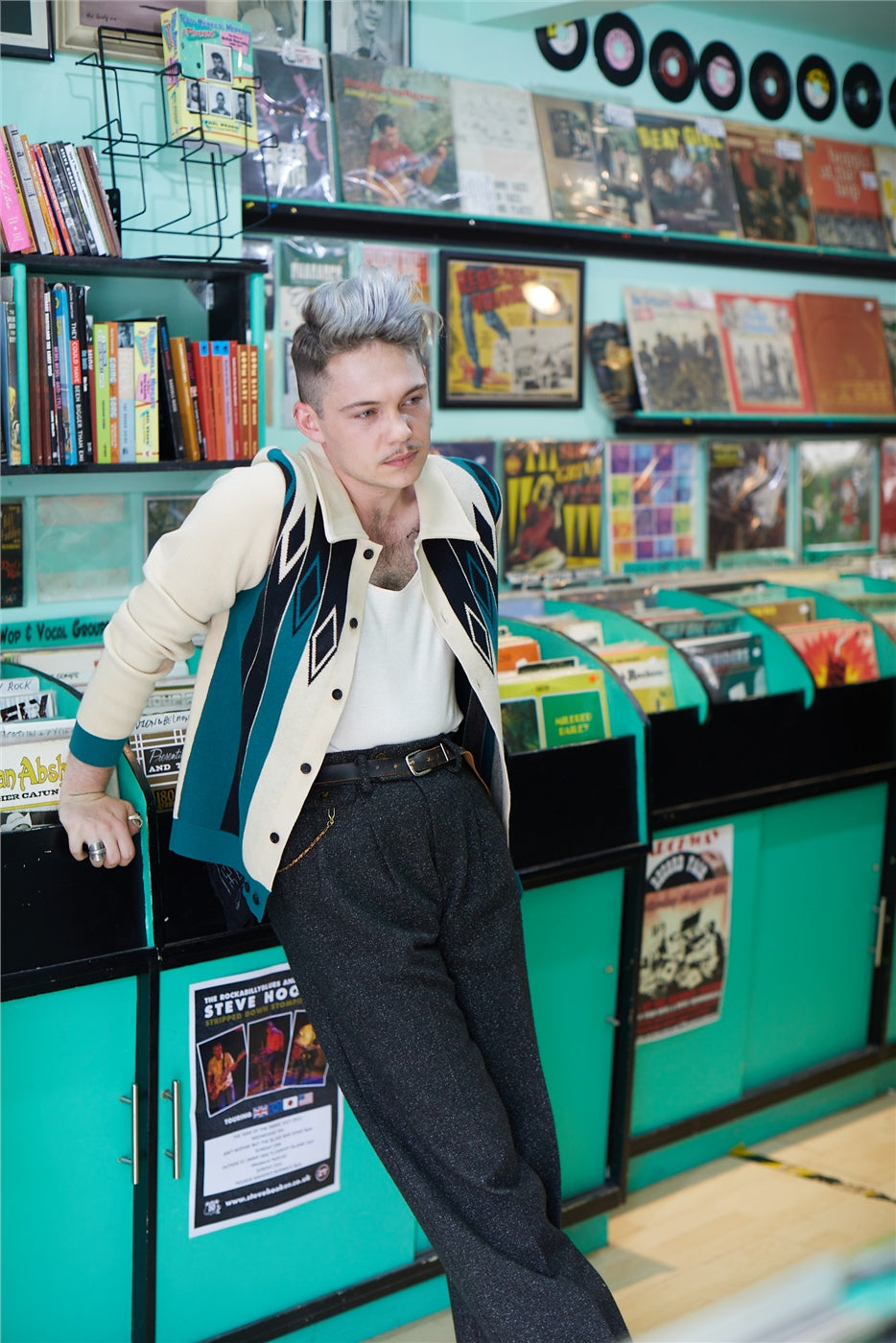 A young man in a retro style records music shop leaing against a counter wearing black jeans, a white t-shirt and a retro cardigan