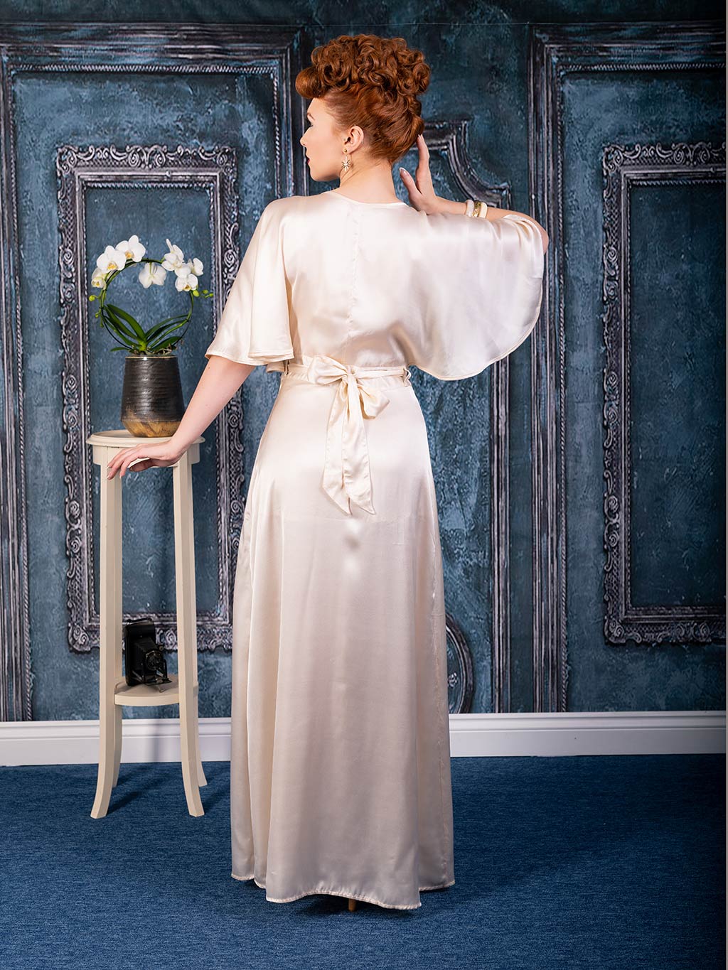  Back view of a model in a 1930s-inspired peach satin dressing gown by What Katie Did, showcasing wide sleeves and a tied sash, styled against a vintage blue and silver panelled wall with an orchid on a side table.