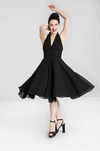 A woman in a vintage-style black dress with a halter neck and full circle skirt, posing with one hand raised and a smile on her face.