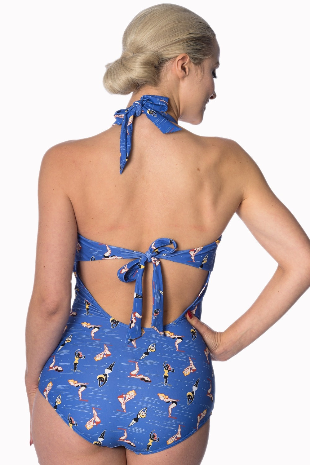 Woman standing with one hand on her hip facing away from the camera  wearing a blue halter neck swimming costume with an all over print of pinup girl cartoon swimmers
