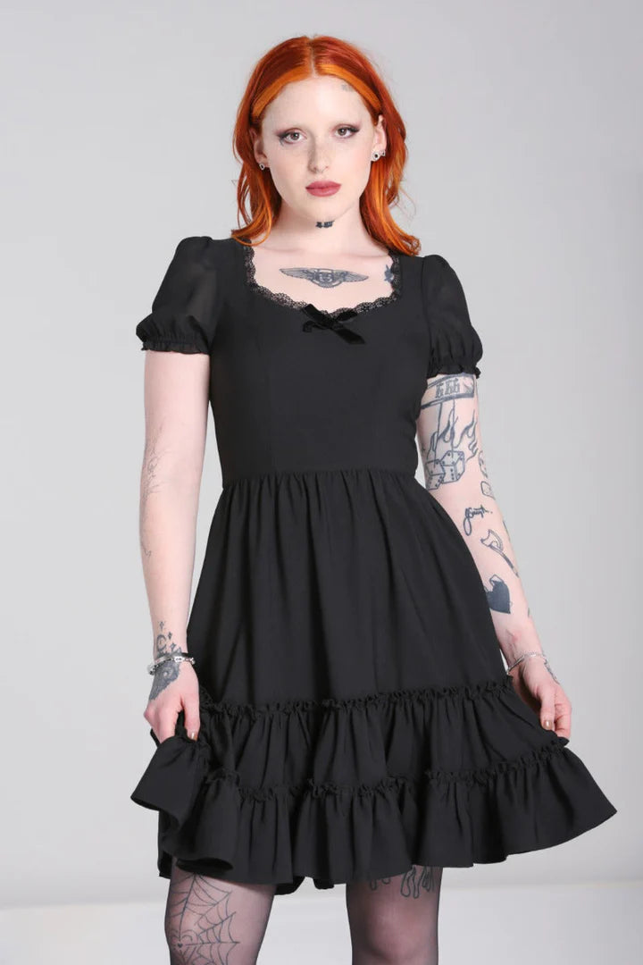Tattooed girl with bright orange hair wearing a black dress standing with one knee bent and looking straight at camera
