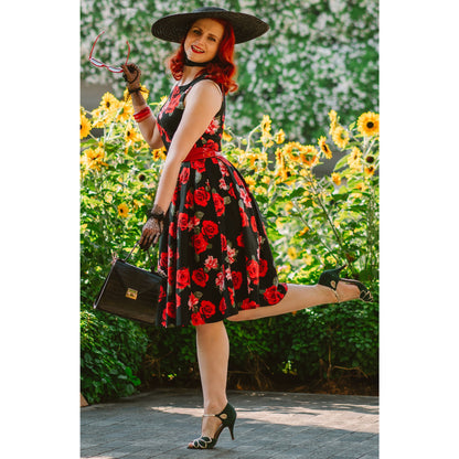 Woman standing in a garden full of sunflowers wearing a sun hat, the Annie Roses Swing dress and stilettos holdinga handbag and a pair of red sunglasses