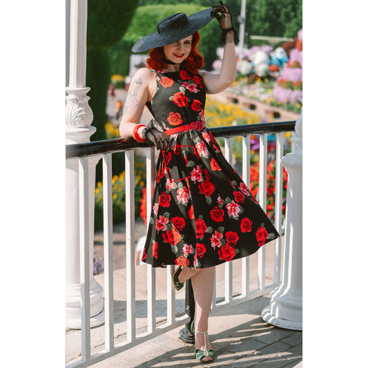 elegant woman leaning against a bandstand rail wearing a wide brimmed black straw hat, black lace gloves, a black swing dress with red rose print and a red belt