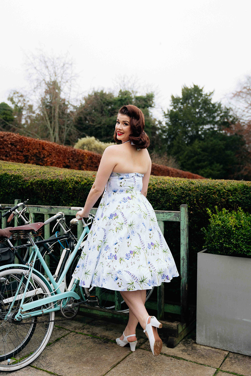 Happy, smiling woman standing next to a bicycle wearing the Bella Swing dress and white heeled shoes