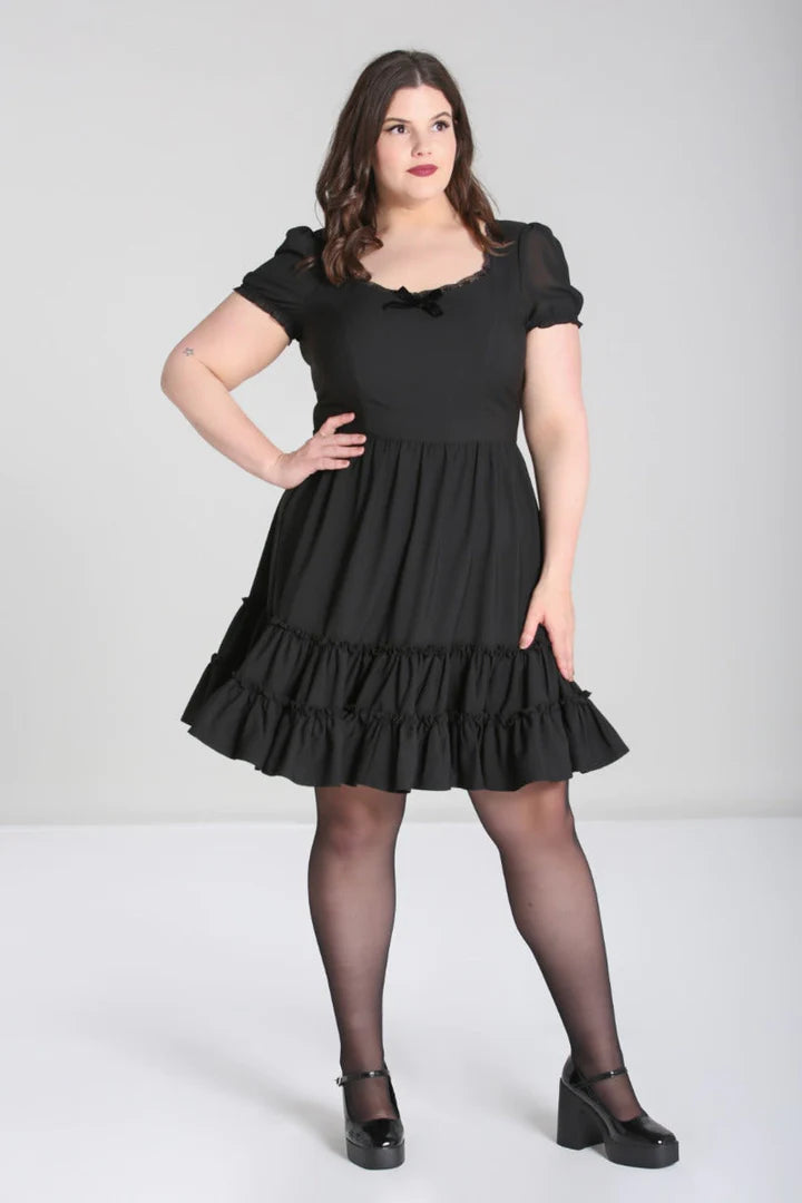 Curvy woman with dark hair and red lipstick stands with a hand on her hip, wearing a black mini dress with short sleeves and frilly hem and black Mary Jane shoes