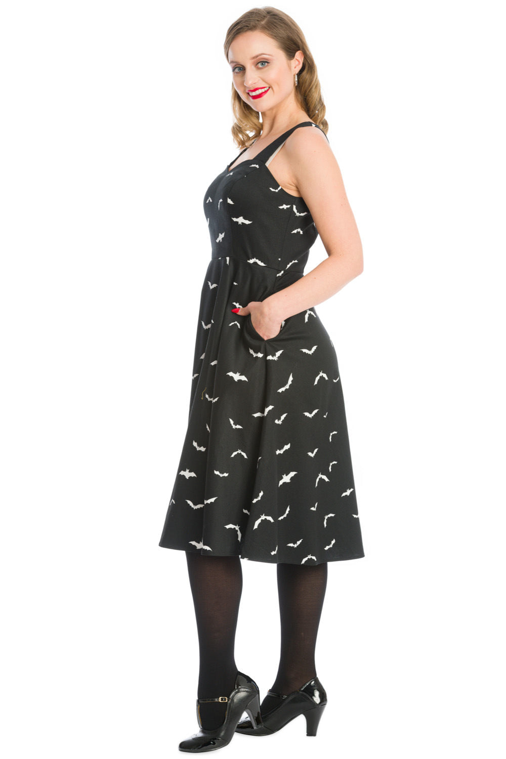 Blonde woman with a warm smile standing gracefully in a stylish black swing dress adorned with an all-over white bat print,  with her hands in the dress pockets 