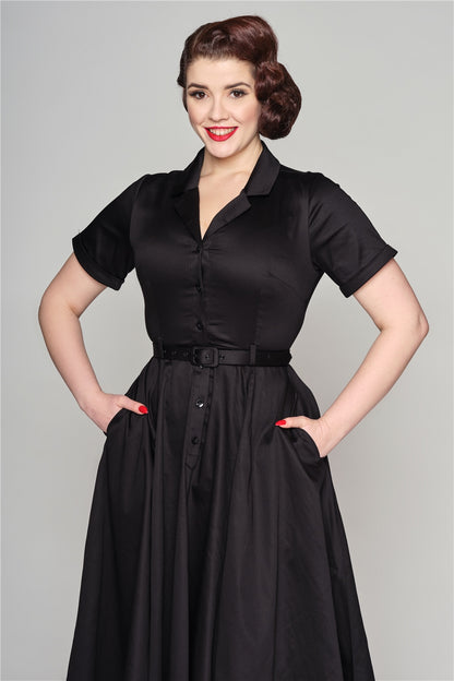Brunette lady smiling, wearing a black dress standing with her hands in her pockets.