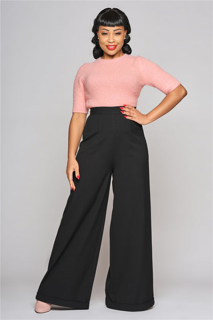 Woman with black hair and a fringe standing with one hand on her hip wearing the Pink Chrissie top and wide leg black trousers