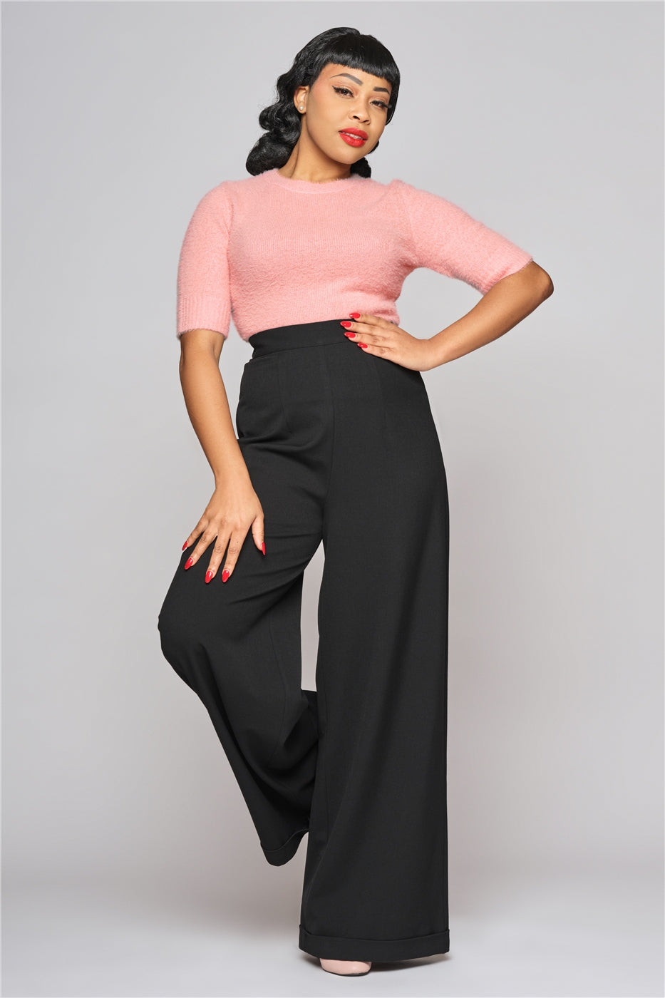 Tall slim woman with red lipstick and nails standing with her hand on her hip and one knee raised wearing the pink Chrissie top and black wide leg trousers