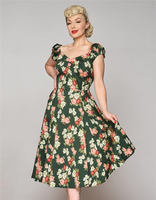 Glamorous blonde woman with a sly smile standing with one hand on her hip and the other holding out the skirt of her green floral print dress 