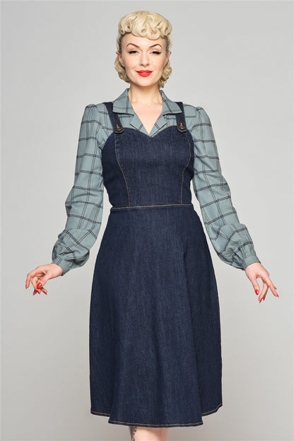 Young blonde woman standing with her arms out by her sides wearing a long sleeved blouse underneath the Jupiter Pinafore Dress by Collectif