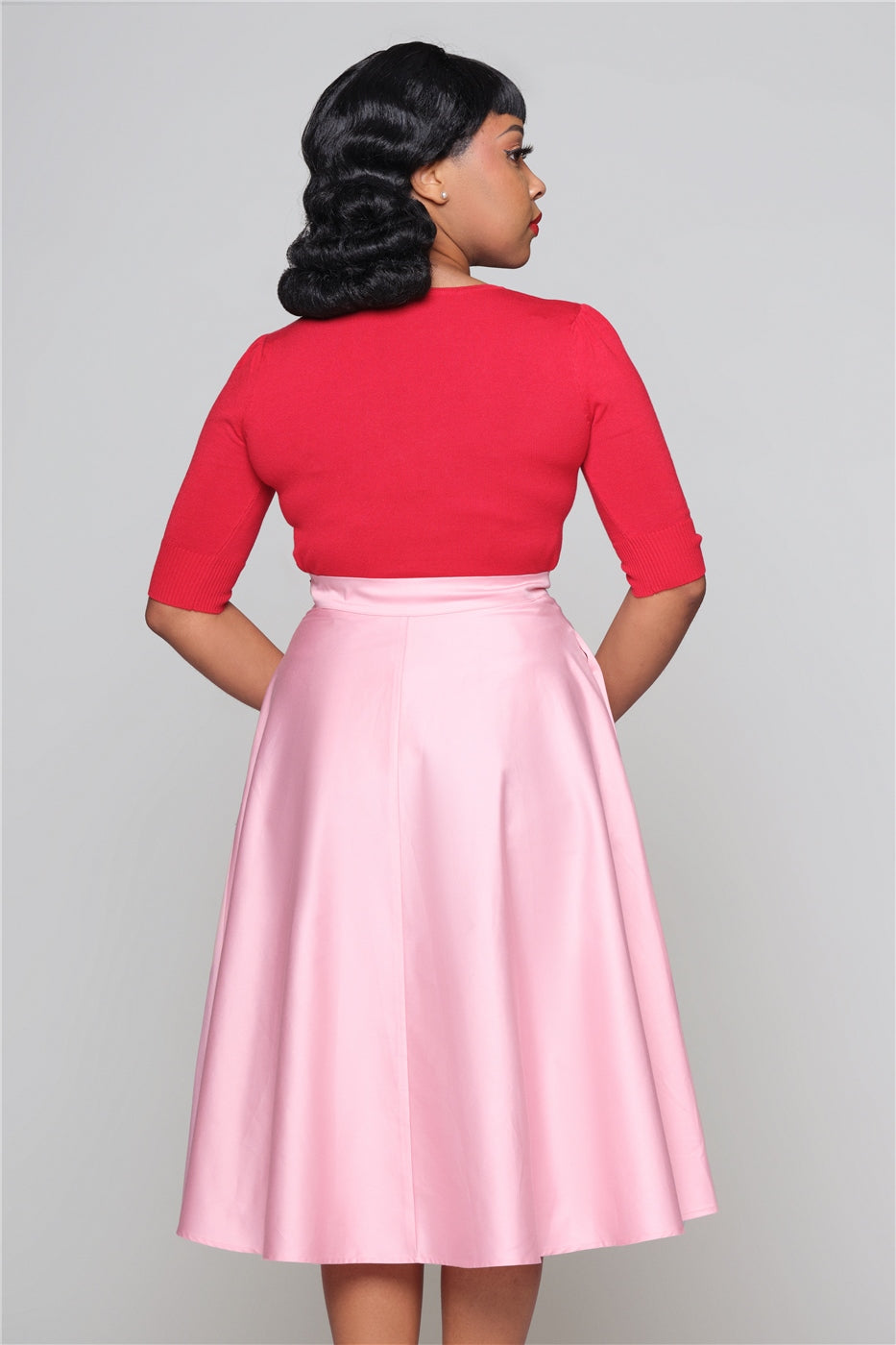 Tall, pretty woman standing straight wearing a red top and pink 50s swing skirt looking over her shoulder