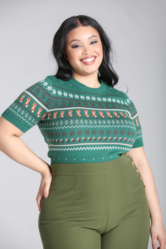 Happy dark haired woman with a radiant smile standing with one hand on her hip wearing the green Vixey knitted short sleeve jumper and green high waisted trousers.