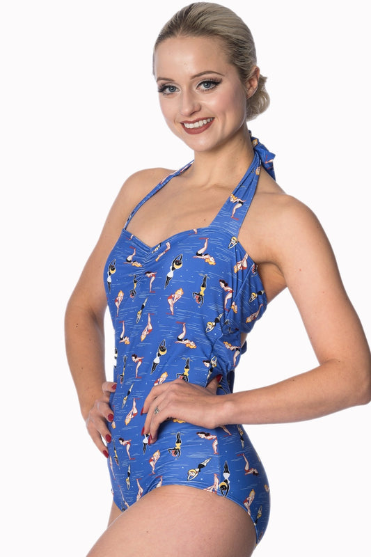 blonde smiling woman standing with her hands on her hips wearing a blue halter neck swimsuit with a print of 50s style pinup girls diving and swimming