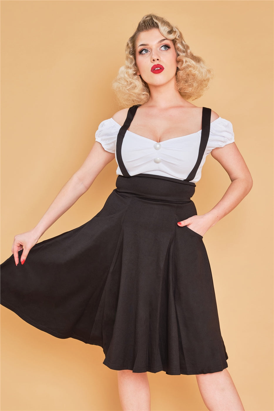 Elegant blonde woman standing against a yellow background wearing a black mid length swing skirt with braces and the white Dolores top 