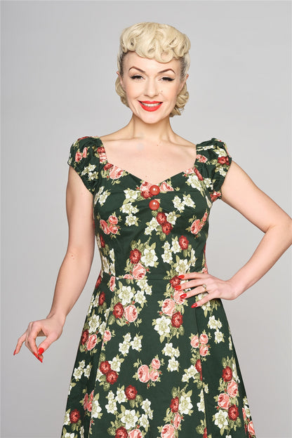 Smiling woman wearing red lipstick and 50s style makeup standing with one hand on her hip. She is wearing a dark green dress with two orange domed buttons at the front , a sweetheart neckline, fitted waist and flared skirt.