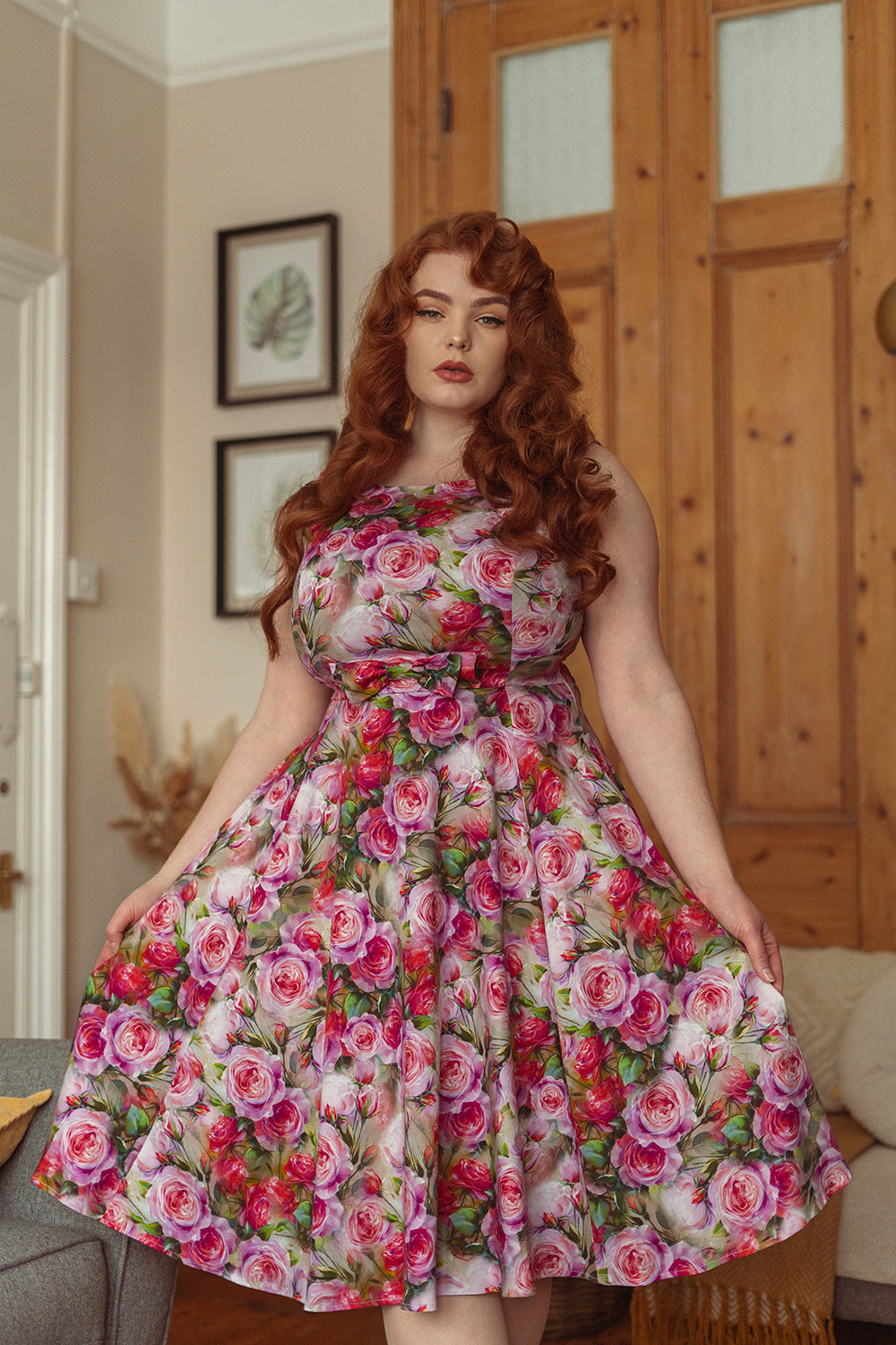 Curvy model with long  red hair standing in her living room holding the skirt of her dress