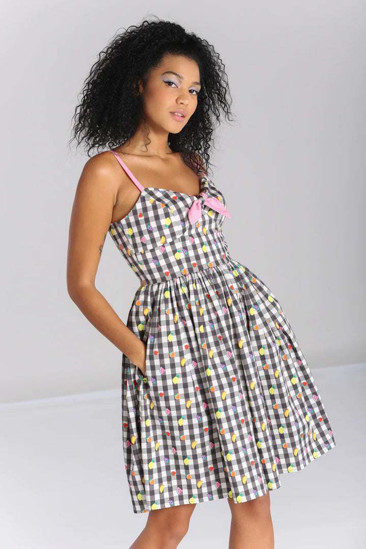 Young woman with curly shoulder length hair standing with her hands in the pockets of her mid length fruit and gingham print dress.
