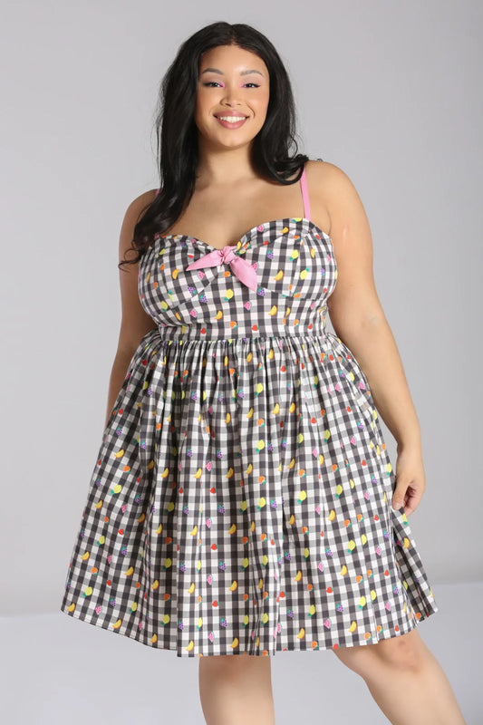 Happy, smiling young woman standing with her arms by her sides wearing a grey and white gingham print dress with pink straps and a tied pink bow at the bust. The dress has a scattered mini colourful fruits pattern all over.