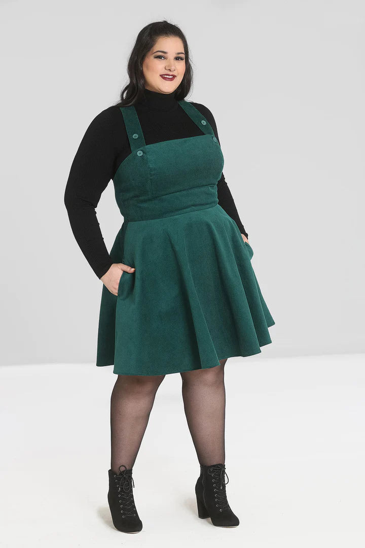 Smiling dark haired woman wearing a high neck black jumper, a green corduroy pinafore dress, tights and black laceup boots