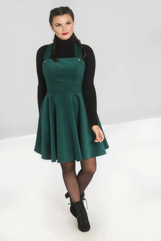 Dark haired woman with plaited hair wearing a black turtleneck jumper and a dark green corduroy pinafore dress with sheer black tights and chunky black boots