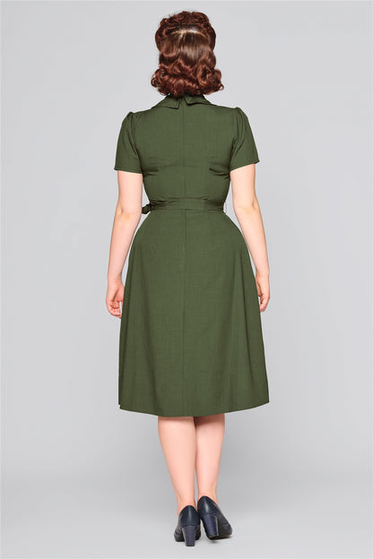 Elegant woman with hair neatly styled and curled wearing a green mid length 40s style dress and black heeled shoes facing away from the camera