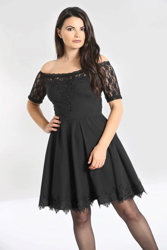 Young woman with long brown hair standing with one hand on her hip. She wears a Bardot black mini dress with short lace sleeves and a lace trim at the bottom of the skirt.