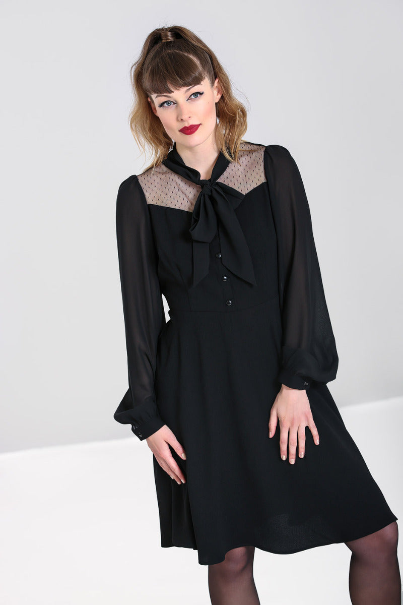 Brunette woman wearing red lipstick standing wearing the Darcia long sleeved black dress with a pussybow tie neck and knee length skirt.