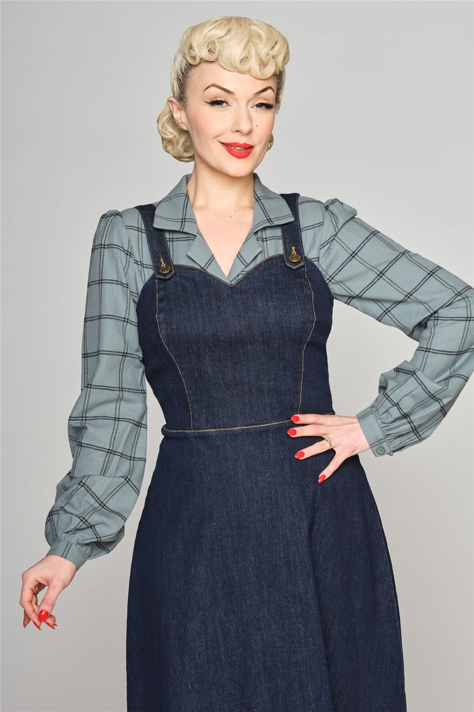 Blonde woman with red lipstick and a demure smile standing with one hand on her hip wearing a blue-grey checked blouse and a denim pinafore dress