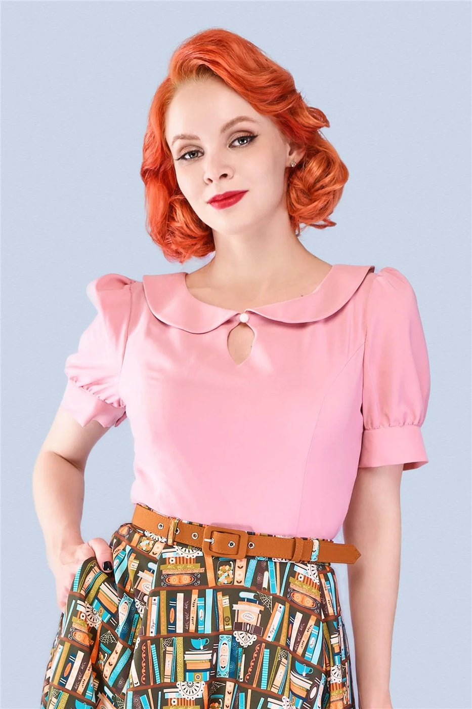Glamorous woman with ginger hair and red lipstick wearing a pink keyhole detail blouse with short puff sleeves and book print skirt standing with one hand in her skirt pocket