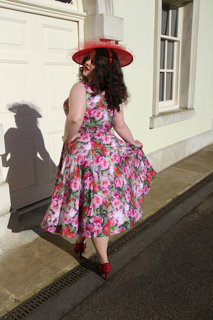 Woman with brown curly hair standing outside wearing a rose print swing dress, red shoes and a red hat.