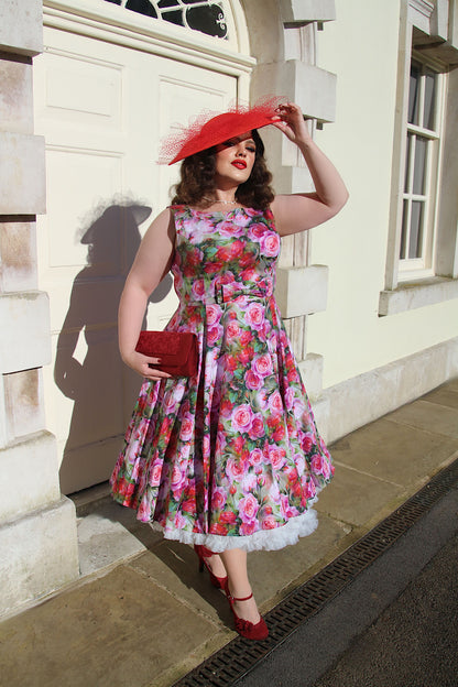 Dark haired sultry woman wearing a red hat and floral dress standing holding a clutch bag in front of a white building 