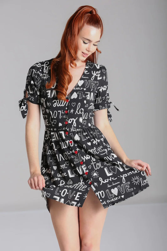 Young girl with long ginger hair tied in a high ponytail pulling the skirt of her mini dress and looking down. The V neck dress has red buttons down the front, short sleeves and white writing all over a black background