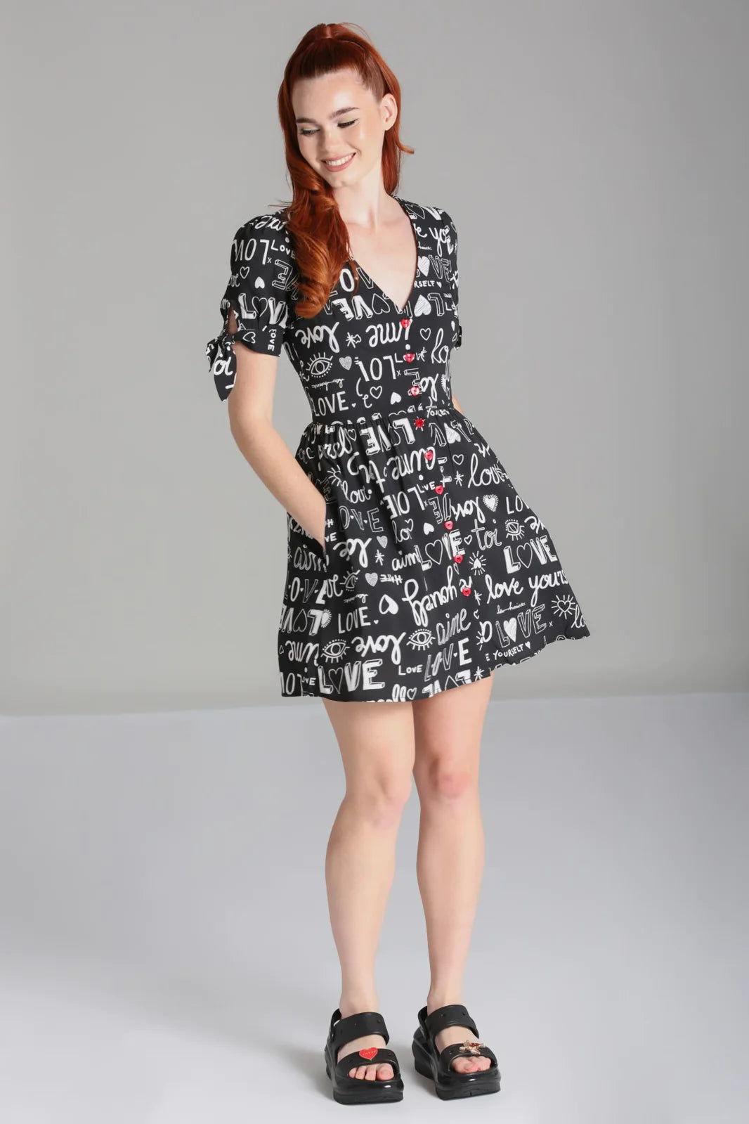 Young girl laughing while standing with her hands in the pockets of her short dress. She is wearing the Love Yourself Mini dress by Hell Bunny and chunky black sandals with a red heart on the toe