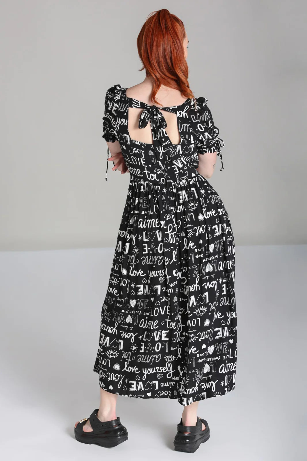 Young woman facing away from the camera showing off the back of the Love Yourself Maxi dress by Hell Bunny which features a tie back detail at the neck.