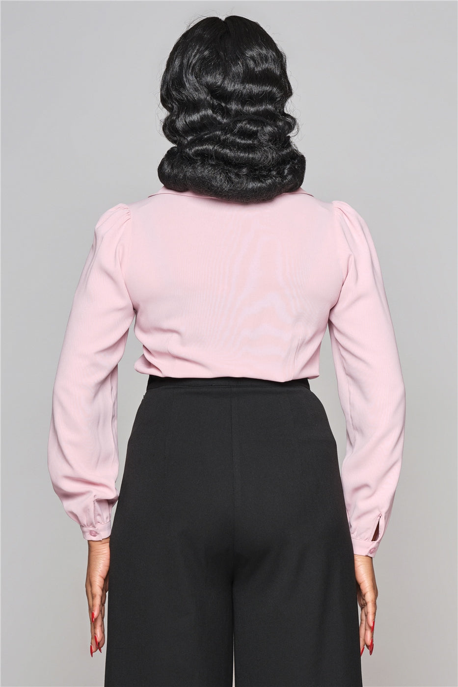 Woman facing away from the camera with black hair in finger waves, wearing a long sleeved pink blouse and high waisted trousers.