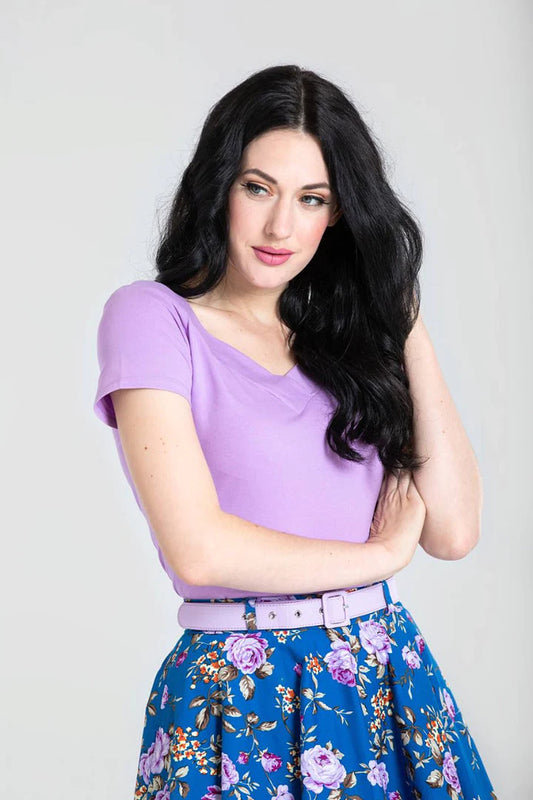 Petite woman with long black hair standing with one arm across her body. She is wearing a light purple Alex top by Hell Bunny and a blue skirt with purple flowers.