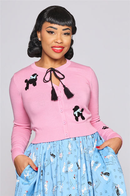Elegant woman with red lipstick standing with her hands in her skirt pockets. She wear a pink cardigan with Fluffy poodle print at the top left bust and bottom right corner and a tie detail black string with tassels.