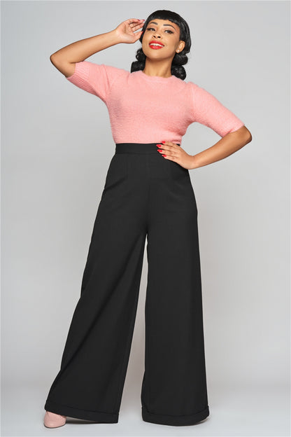 Young elegant woman standing with one hand on her hip and the other to her face wearing the pink fluffy Chrissie knit top and wide leg black trousers