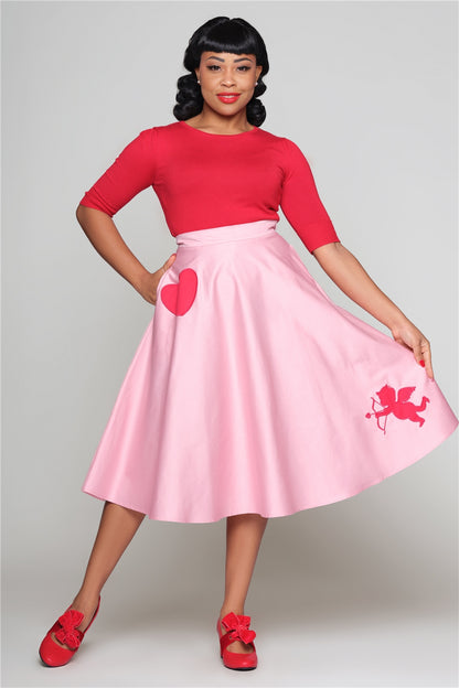 Vintage styled woman with black hair wearing a red top and shoes and a pink 50s style swing skirt with a red heart at the top left pocket and a cupid motif at the bottom right