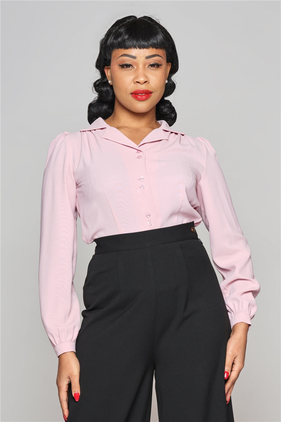 Pretty lady with a vintage shoulder length hair style  standing with her hands by her sides smiling softly,  wearing a beautiful pink blouse and high waisted black trousers.