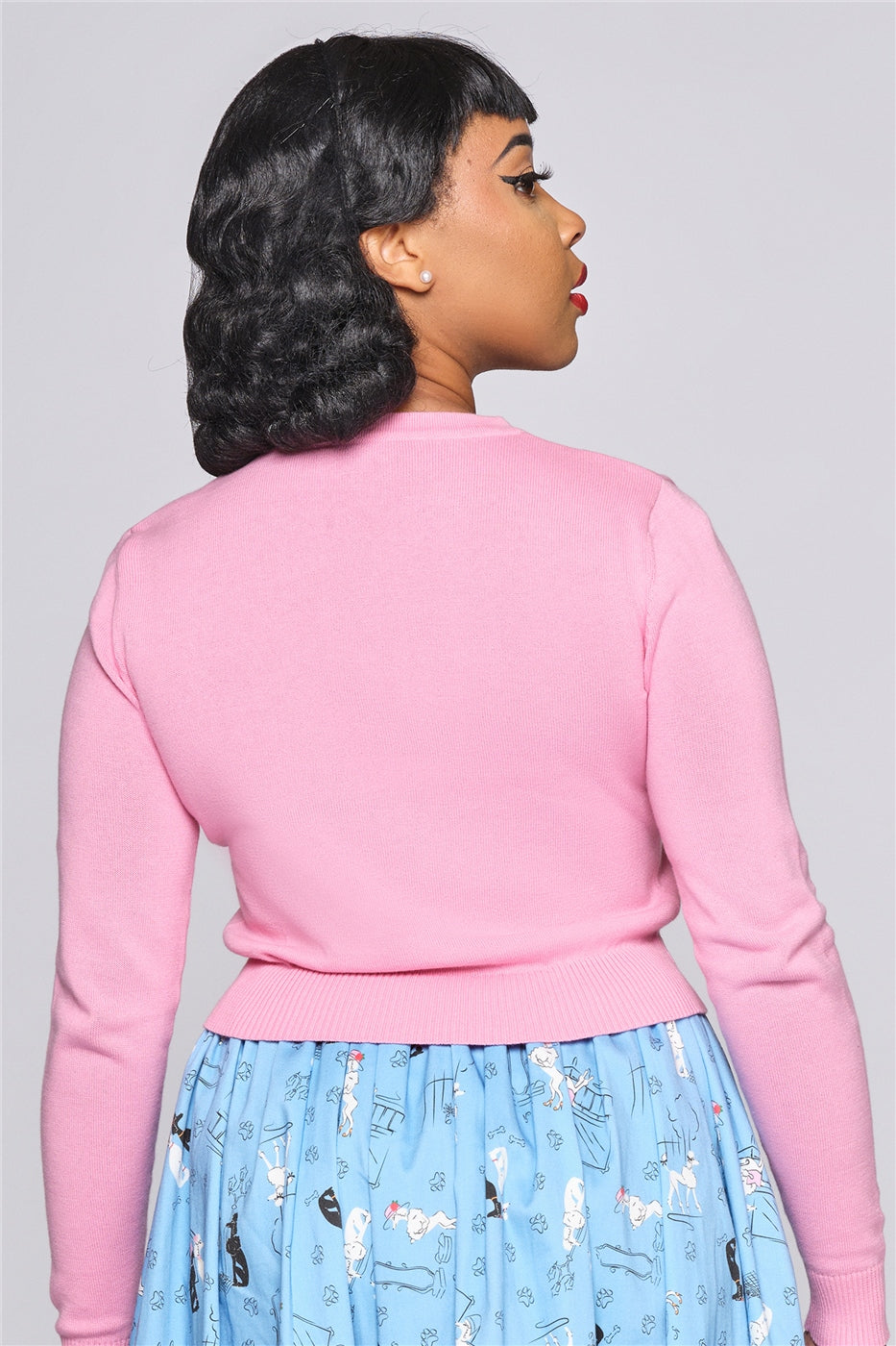 Tall model with short black hair standing facing away from the camera showing the plain pink back of the Char Poodle Cardigan