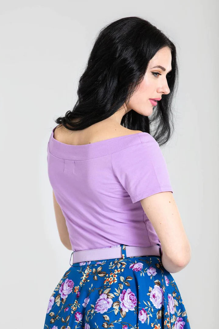 Dark haired young woman standing facing away from the camera. She is wearing a short sleeved plain t shirt top and a blue floral skirt with a lavender coloured belt.
