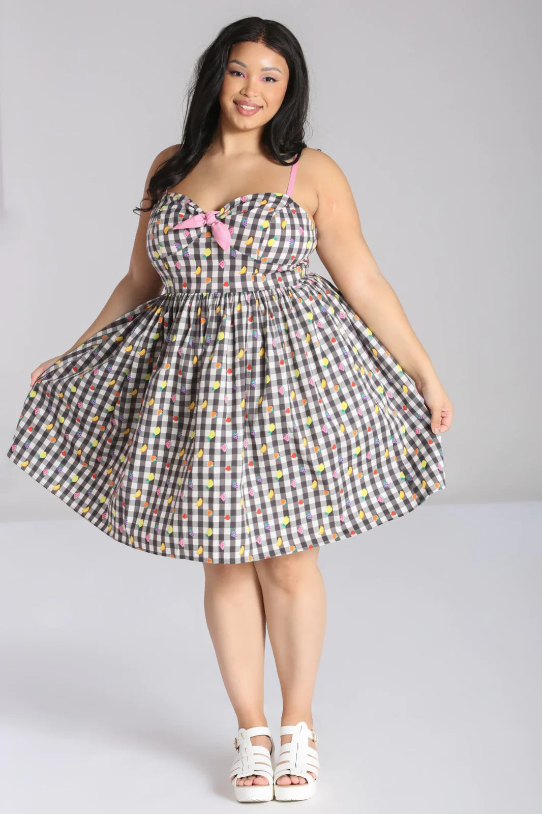 Dark haired woman with a radiant smile standing holding her skirt out at the sides. She is wearing the Fruity Lou Gingham dress by Hell Bunny and chunky white sandals.