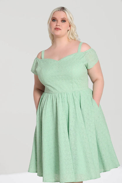 Elegant blonde woman wearing natural makeup standing with her hands in her pockets wearing a mint green broderie anglaise dress with cold shoulder detail