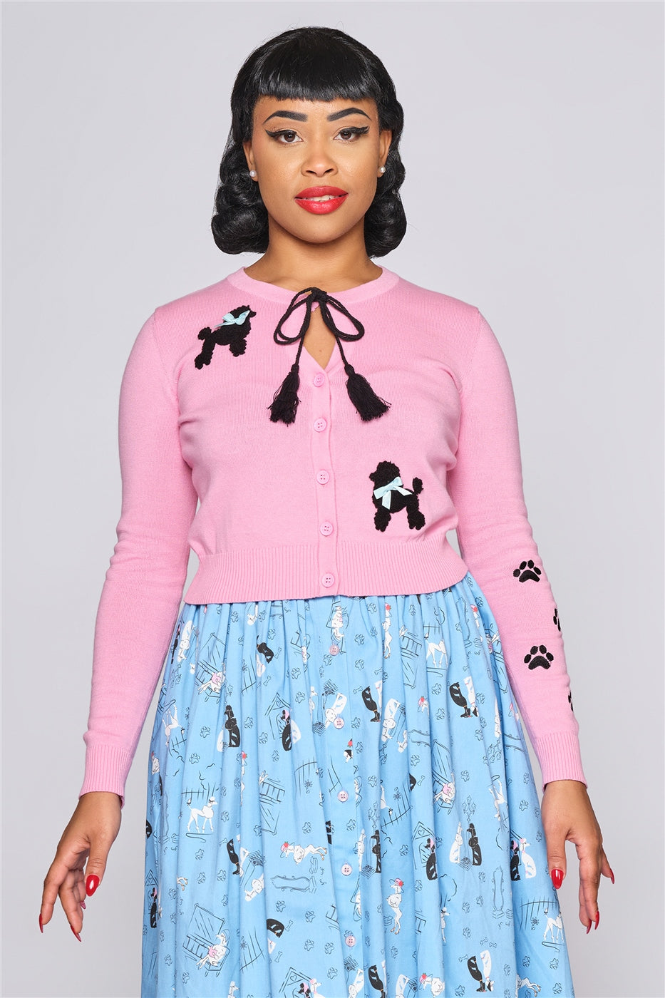 Glamorous woman with short curled black hair wearing a blue poodle print skirt and pink cardigan with Poodle motif