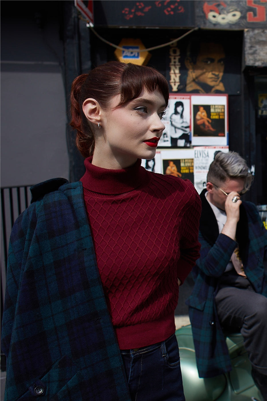 Brunette vintage styled model wearing a burgundy sweater, high waisted vintage jeans and a blue/green tartan coat around her shoulders standing outside a shop 